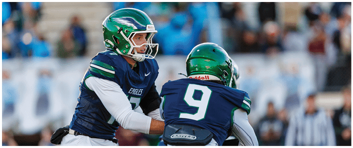 Two people wearing green helmets and navy blue football uniforms are engaged in a play on a football field with blurred spectators in the background. Description generated by AI