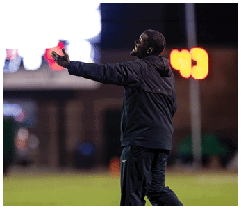 A person wearing a dark jacket and pants is standing on a field with arms outstretched, appearing to be shouting or calling out. Description generated by AI