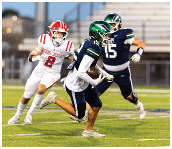 A person in a green helmet and navy uniform is running with a football while two people in white and navy uniforms chase on a grassy football field. Description generated by AI