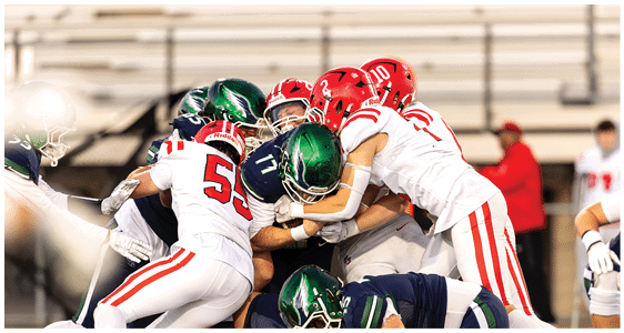 Several football players in red and white and blue and green uniforms are engaged in a close tackle on the field with empty bleachers in the background. Description generated by AI