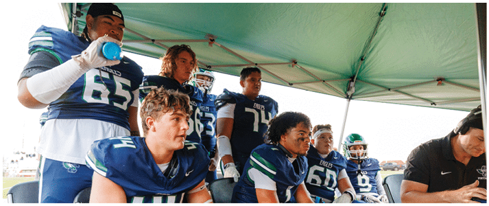 A group of people wearing football uniforms are sitting and standing under a canopy, appearing focused and attentive. Description generated by AI