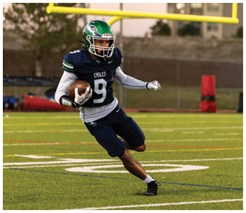 A person wearing a green helmet and dark blue football uniform with the number 9 is running on a football field while holding a football. Description generated by AI