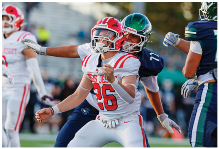 A person wearing a white and red football uniform is being closely defended by two people in dark blue and green football uniforms during a game. Description generated by AI