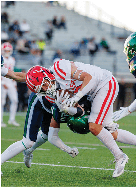 A person in a white and red football uniform is tackling another person in a green and blue football uniform on a grassy field with blurred spectators in the background. Description generated by AI