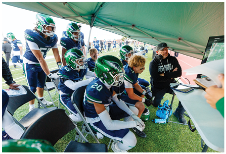 People in football uniforms and a coach are seated and standing under a tent on the sidelines of a football field, watching a screen and discussing strategy. Description generated by AI
