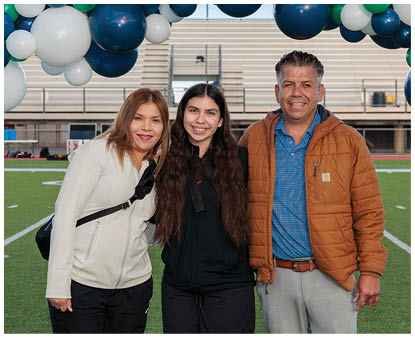 Three people standing closely together smiling under a balloon arch on a sports field with bleachers in the background. Description generated by AI