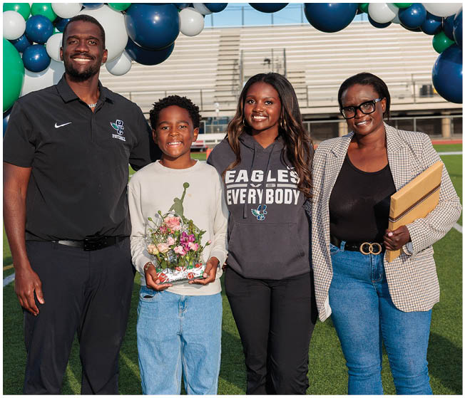 A cluster of green, navy blue, and white balloons is in the foreground on a sports field with people in sports uniforms and bleachers in the background. Description generated by AI