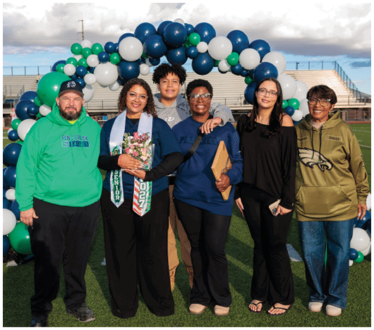 A group of people stands together outdoors under a balloon arch, with one person wearing a senior sash and holding flowers while others smile and pose closely. Description generated by AI