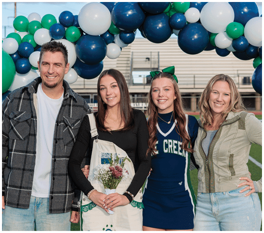 A person wearing a plaid shirt smiles in front of a large cluster of green, white, and blue balloons with bleachers and a partly cloudy sky in the background. Description generated by AI