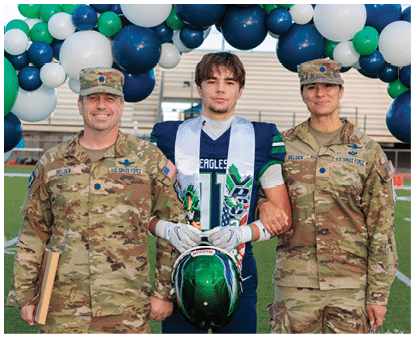 A person wearing a football uniform and a white celebratory sash stands between two people in military uniforms, all smiling and posing outdoors. Description generated by AI