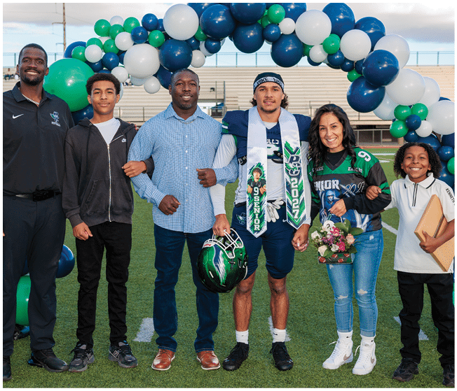A group of six people stand together on a sports field under a balloon arch, with one person wearing a football uniform and holding a helmet, and another holding flowers. Description generated by AI