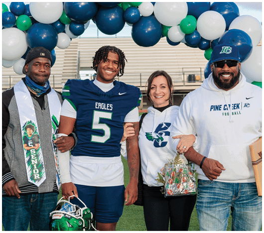 Four people stand arm in arm under a balloon arch on a sports field, with one person wearing a football uniform and holding a helmet, while the others wear casual clothing including a senior sash and team apparel. Description generated by AI