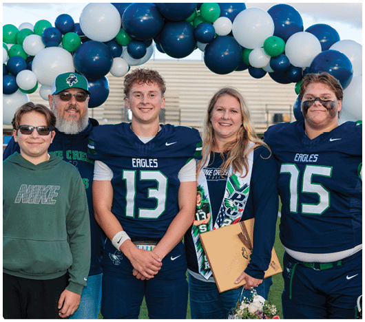 Five people stand smiling under a balloon arch with two wearing football uniforms numbered 13 and 15, one holding flowers and a folder, and the others dressed casually. Description generated by AI