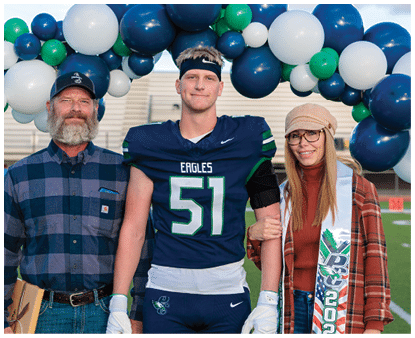 Three people stand on a sports field under an arch of blue, white, and green balloons, with one person in a football uniform holding a helmet and the other two dressed casually, one wearing a sash with “2027" on it. Description generated by AI