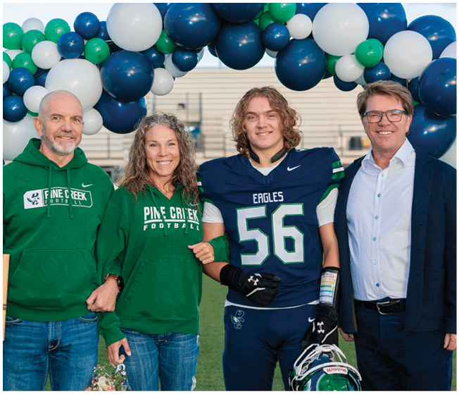 Four people stand together smiling on a sports field under a balloon arch, with one person wearing a football uniform and holding a helmet, two wearing matching green hoodies with “Pine Creek Football" text, and one in a white shirt and glasses. Description generated by AI
