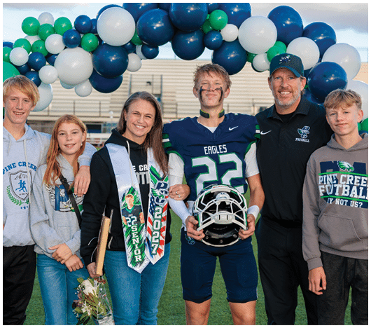 A group of six people stand together on a sports field under a balloon arch, with one person in a football uniform holding a helmet and others wearing casual clothing and smiling. Description generated by AI