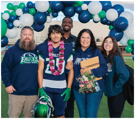 Five people stand together on a sports field under an arch of blue, white, and green balloons, with one person wearing a football uniform and a flower lei, holding a helmet, and others smiling and holding flowers and a folder. Description generated by AI
