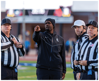 A person wearing a black jacket and beanie is pointing while standing next to three people in black and white striped referee uniforms with headsets on a sports field. Description generated by AI