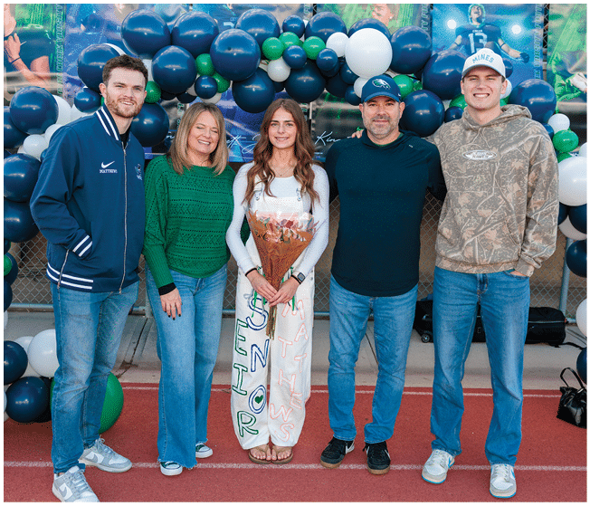Five people stand on a track in front of a balloon arch with one person in the center holding flowers and wearing a white outfit decorated with colorful writing. Description generated by AI
