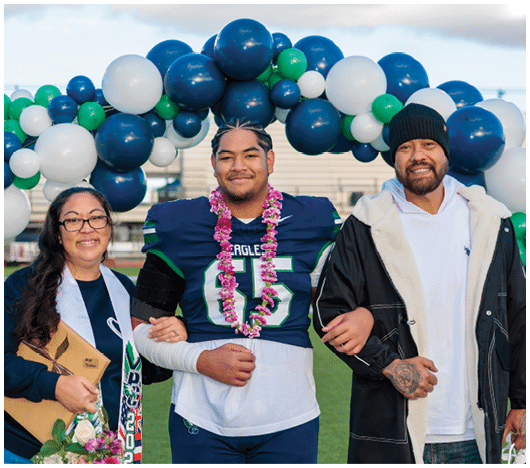 Three people stand arm in arm on a sports field under a balloon arch, with the person in the center wearing a football uniform and a flower lei, smiling. Description generated by AI