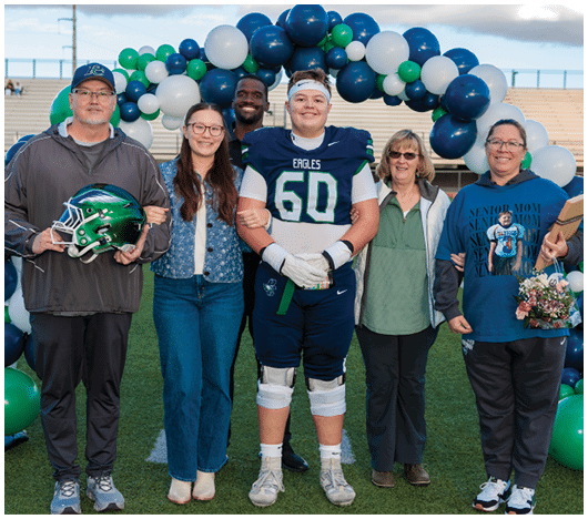 A group of six people stand on a football field under a balloon arch, with one person in a football uniform holding a belt and others dressed casually, some smiling and one holding a football helmet. Description generated by AI