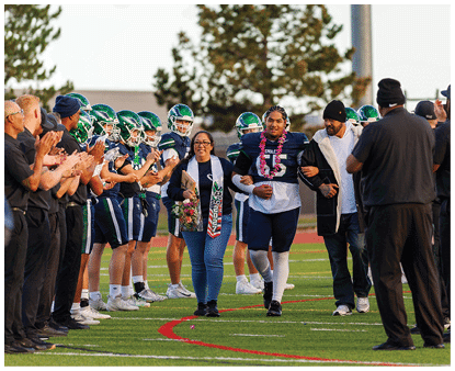 A person wearing a football uniform and a flower lei is walking arm-in-arm with two people, one holding flowers and a sash, while football players in helmets stand in a line clapping on a field. Description generated by AI