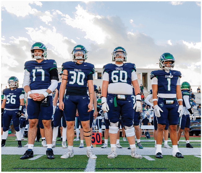 Four people in football uniforms and helmets stand side by side on a field with a partly cloudy sky in the background. Description generated by AI
