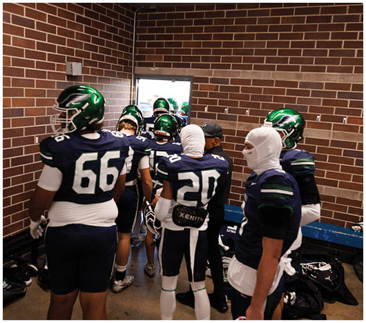 A group of people wearing green helmets and dark sports uniforms are standing in a brick-walled hallway, preparing to enter a brightly lit area. Description generated by AI