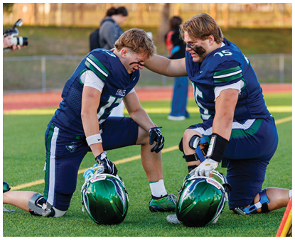 Two people in football uniforms kneel on a field with helmets on the ground while one person places a hand on the other's shoulder. Description generated by AI