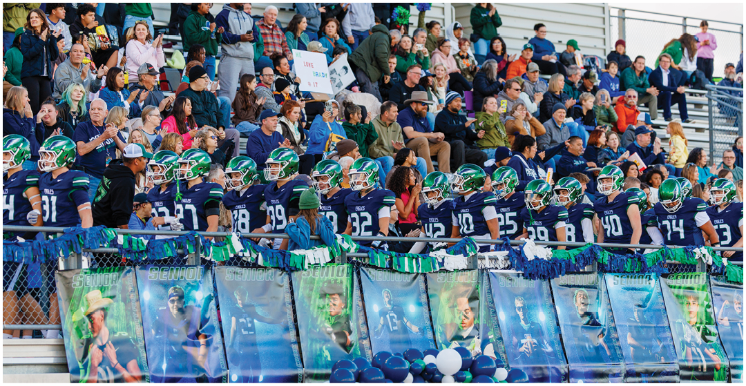 A crowd of people is seated in bleachers watching a football game while players in helmets and uniforms stand in front of banners and balloons. Description generated by AI