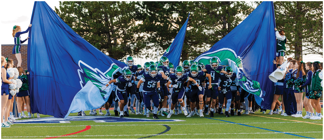 A group of football players in dark uniforms with helmets are running onto a field through large blue banners while cheerleaders stand on the sidelines. Description generated by AI