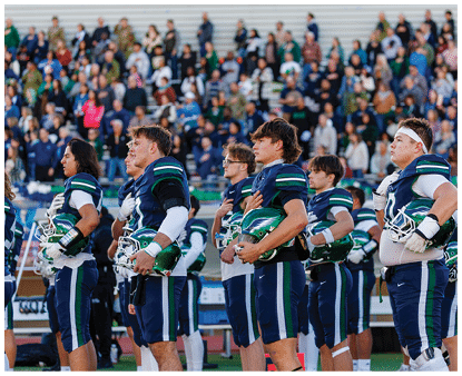 A group of people in football uniforms stand in a line on a field with their hands over their hearts, holding helmets, while a crowd watches in the background. Description generated by AI