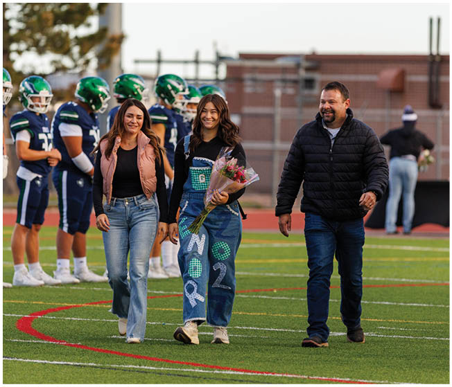Three people walk together on a sports field with one person holding a bouquet of flowers and wearing decorated overalls while football players stand in the background. Description generated by AI