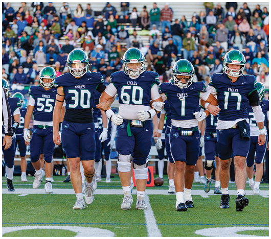 Five people in football uniforms with green helmets walk arm in arm on a field with a crowd in the background. Description generated by AI