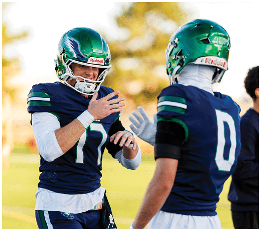 Two people wearing green helmets and navy blue football uniforms are interacting on a field with a blurred background. Description generated by AI