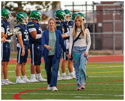 People in sports uniforms and casual clothing stand on a football field with a chain-link fence and trees in the background. Description generated by AI