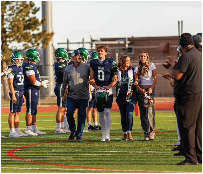 A group of people on a football field includes a person in a football uniform walking arm-in-arm with three others while several teammates stand in the background. Description generated by AI