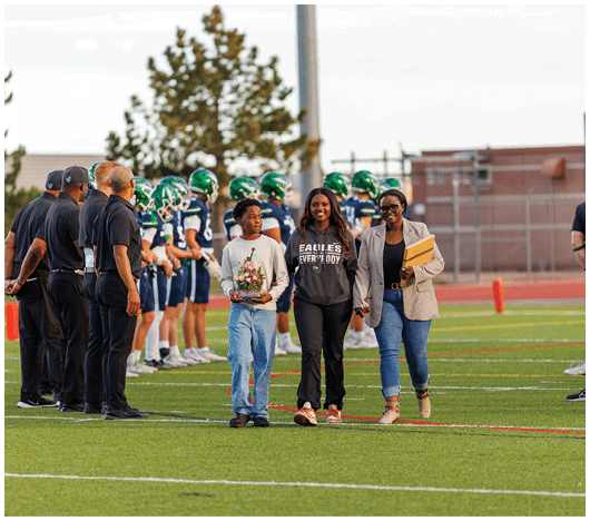 People are standing on a football field with a group of players in uniform lined up in the background while three people walk together in the foreground, one holding flowers and another holding a folder. Description generated by AI