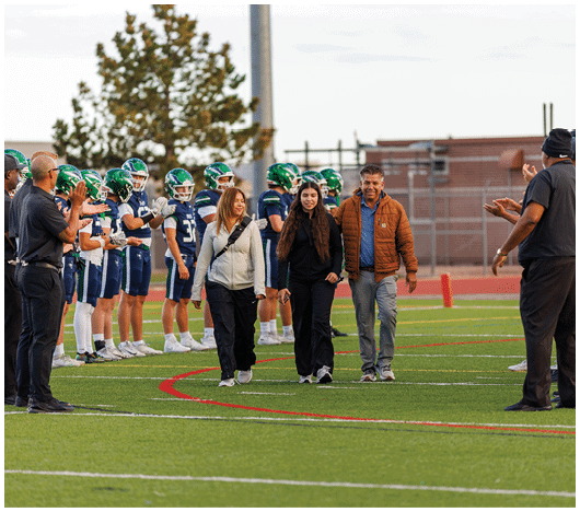 Three adults walk together on a football field while players in green helmets and blue uniforms stand in lines on either side, and other adults in dark clothing applaud. Description generated by AI
