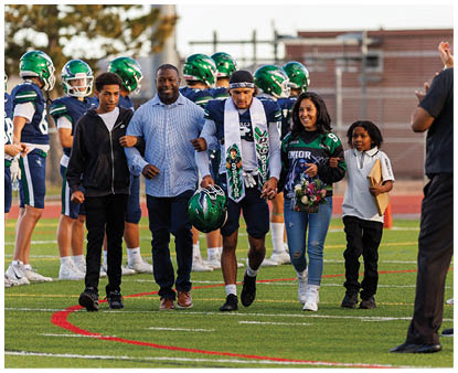 A group of people walk arm in arm on a football field with one person in a football uniform holding a helmet and wearing a senior sash, while others wear casual clothing and one holds flowers. Description generated by AI