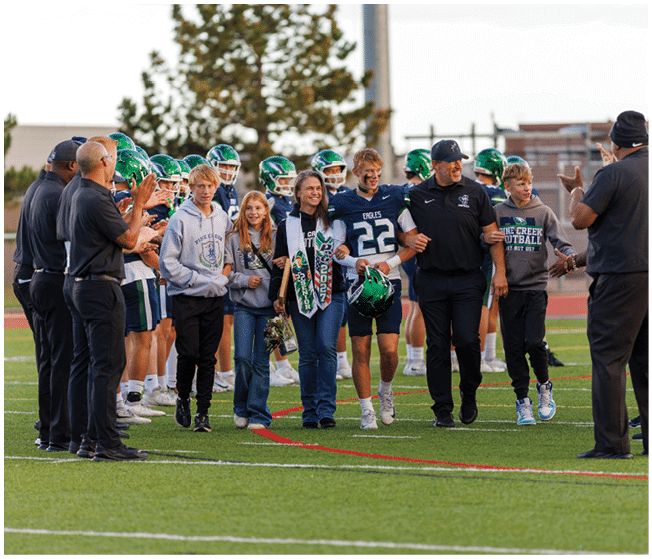 A group of people including athletes in football uniforms and others in casual clothing walk arm in arm on a football field during what appears to be a celebratory or ceremonial event. Description generated by AI
