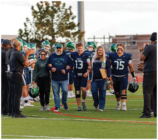 People in football uniforms and casual clothing walk together on a field while others in football gear stand in lines clapping. Description generated by AI