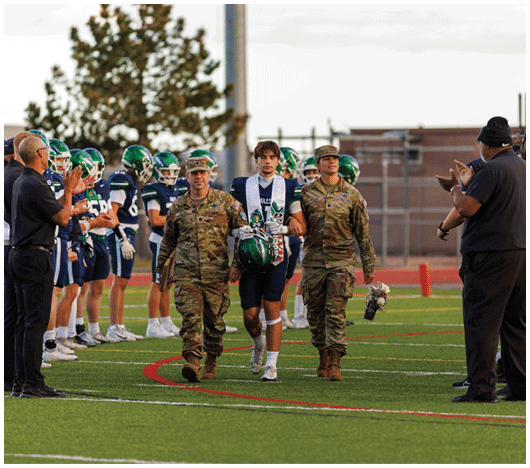 Two adults in military uniforms walk alongside a person in a football uniform holding a helmet on a football field while people on the sidelines clap. Description generated by AI