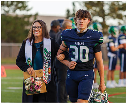 An adult wearing a dark coat and a colorful sash holds a bouquet and a folder while walking arm-in-arm with a person in a football uniform holding a helmet on a sports field. Description generated by AI