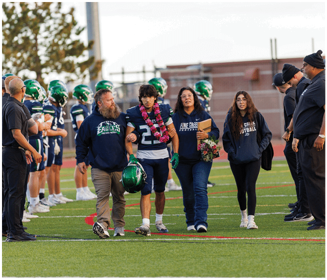 A person wearing a football uniform with a lei around their neck is walking arm-in-arm with three other people on a football field while a line of players in helmets and uniforms stand in the background. Description generated by AI