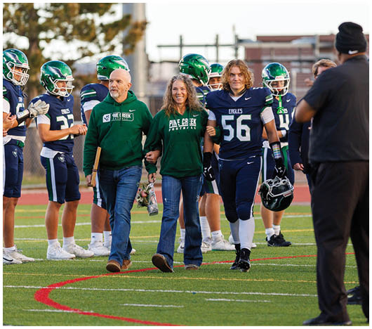 A person in a football uniform walks arm-in-arm with two adults wearing matching green sweatshirts while a line of football players in helmets and uniforms stand on the field clapping. Description generated by AI