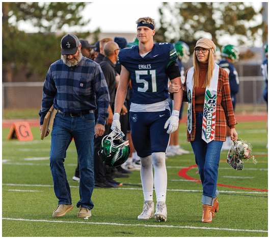An adult in a football uniform holding a helmet is walking arm-in-arm with two adults on a football field, one holding flowers and wearing a scarf with a year on it. Description generated by AI