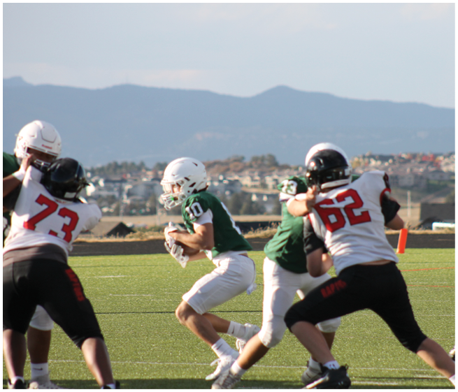 Four people in football uniforms are engaged in a play on a grassy field with mountains and buildings in the background. Description generated by AI