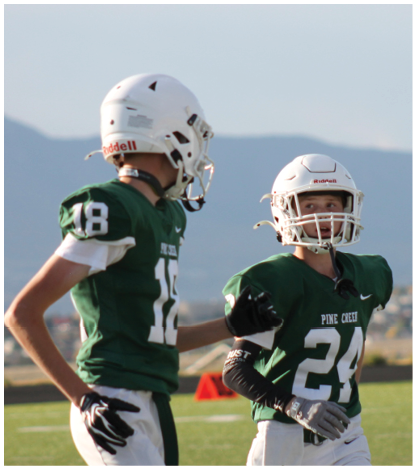 Three people wearing green football uniforms and white helmets stand on a field with mountains in the background. Description generated by AI