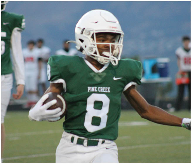 A person wearing a green football uniform with the number 8 is running with a football while another person in a green uniform with the number 65 stands in the background on a football field. Description generated by AI